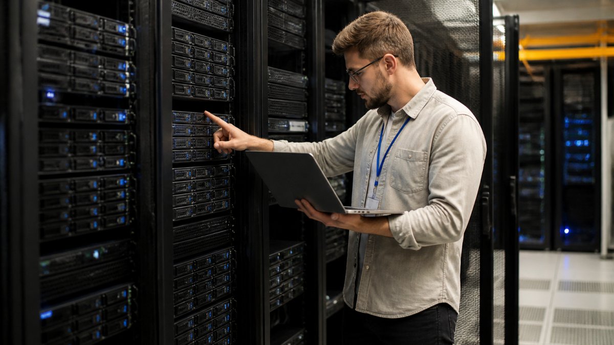 A data center technician in a server room inspecting rack-mounted servers with a laptop, rows of blinking server units in the background