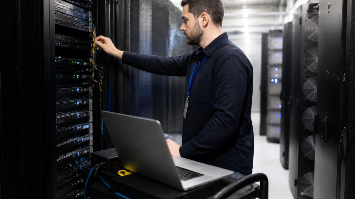 A system administrator in a data center aisle inspecting a rack of dedicated servers, with a laptop open on a rolling cart showing server monitoring dashboards
